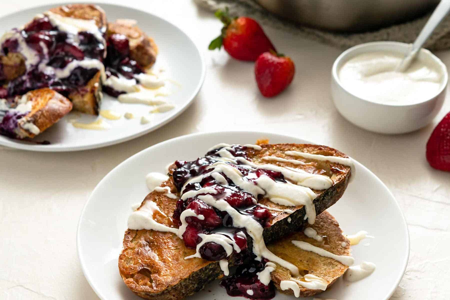Image of two plates of berry sourdough French toast on an off-white background with cream sauce
