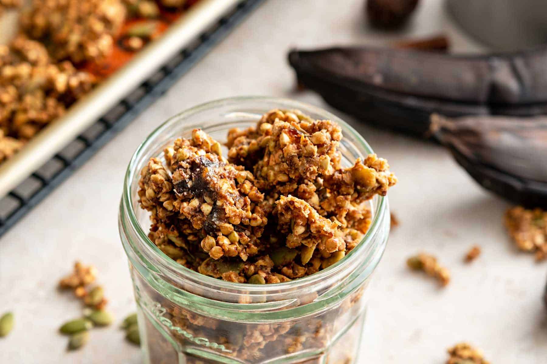 Jar of buckwheat granola next to ripe bananas and baking sheet
