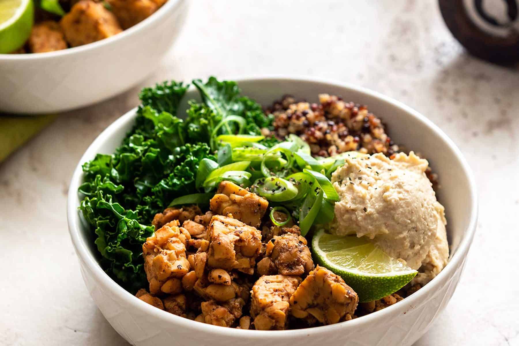 Two white bowls with kale, tempeh, quinoa, and bean dip on a marble background