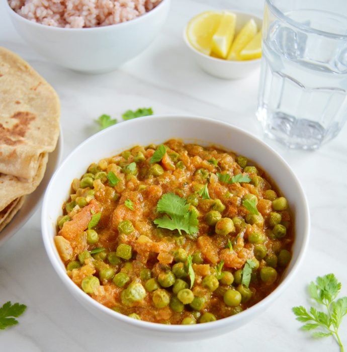 green peas masala in a white bowl with chapatis, brown rice, lemon slices and a glass of water.