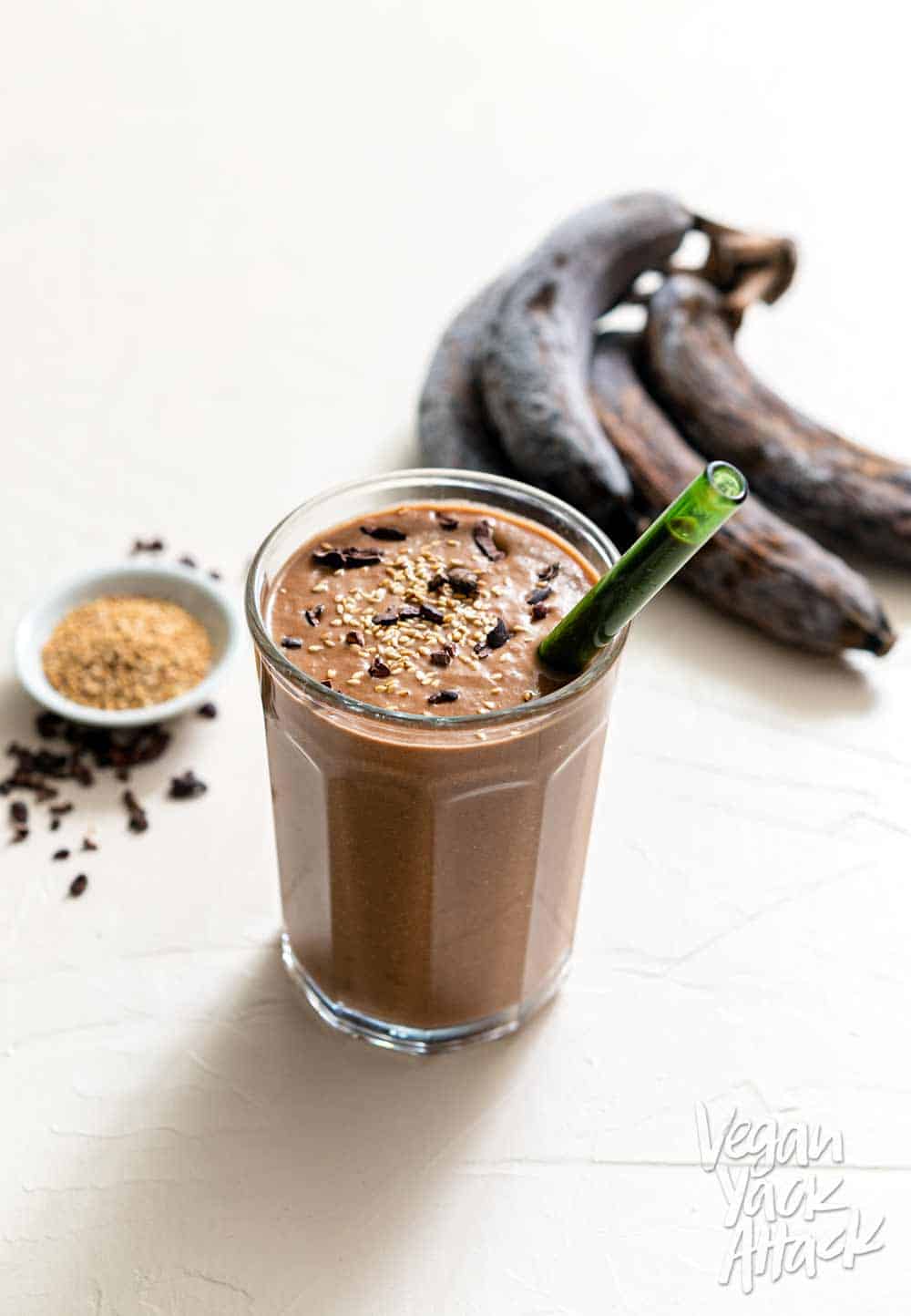 One large glass filled with chocolate sesame smoothie, with seeds, nibs, and bananas on the side