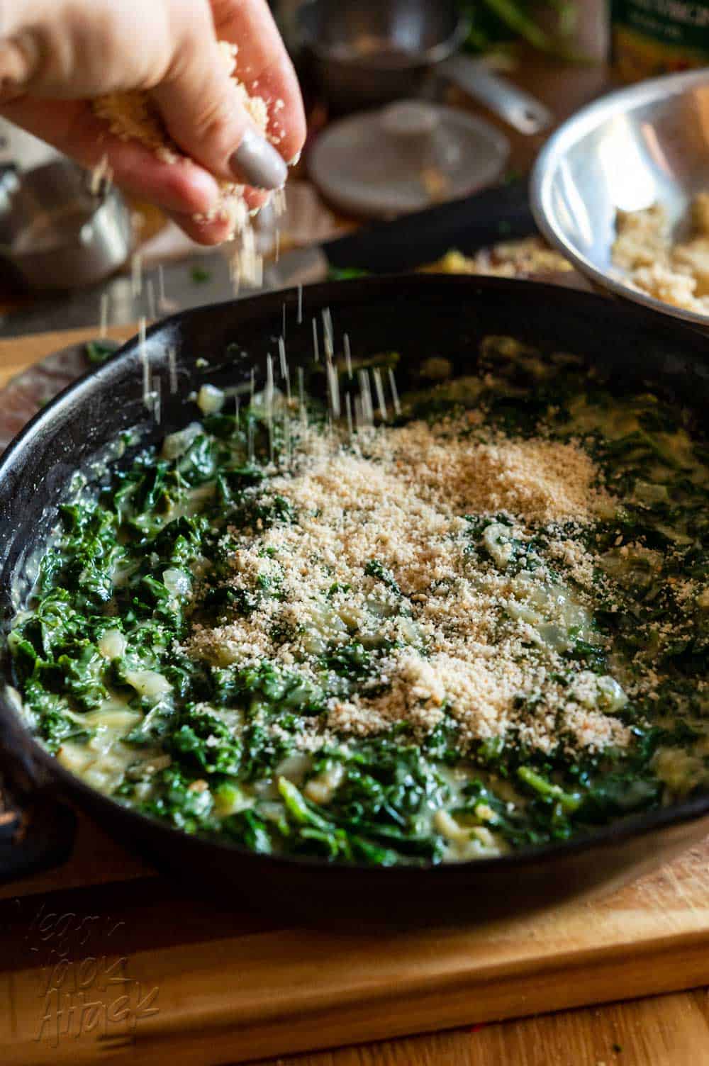Hand sprinkling bread crumbs over creamed kale in a skillet
