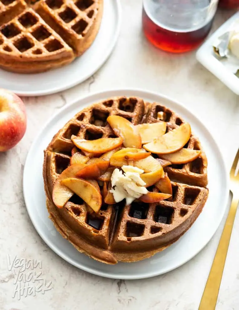 2 large, round, Belgian waffles on white plates, topped with apples and with syrup on the side