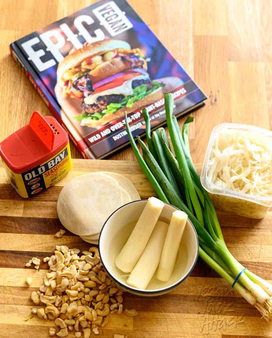 Hearts of palm in a bowl, next to green onions, gyoza wrappers, old bay seasoning, cashew pieces, and sauerkraut, on a cutting board, with a copy of the Epic Vegan Cookbook behind it.