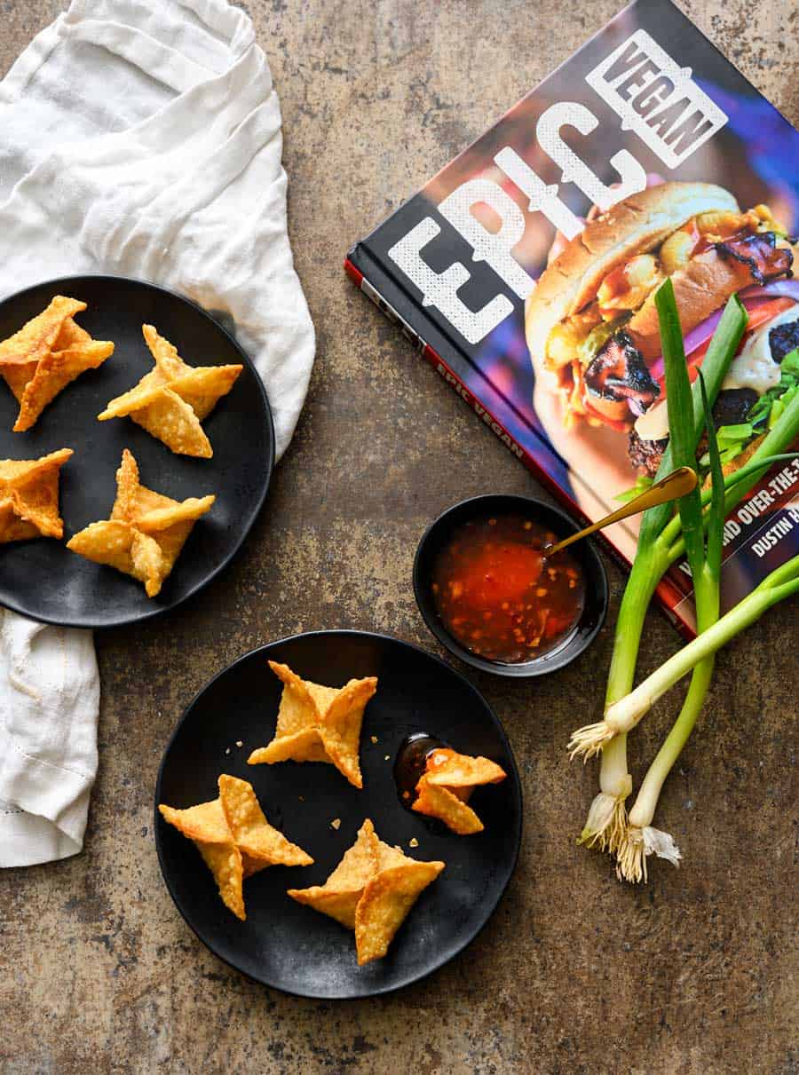 Vegan Crab Rangoon on black plates with a side of sweet chili sauce, whole green onions, and a white linen on a grey background, with the Epic Vegan Cookbook.