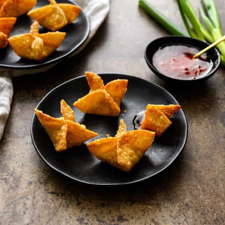 Vegan Crab Rangoon on black plates with a side of sweet chili sauce, whole green onions, and a white linen on a great background