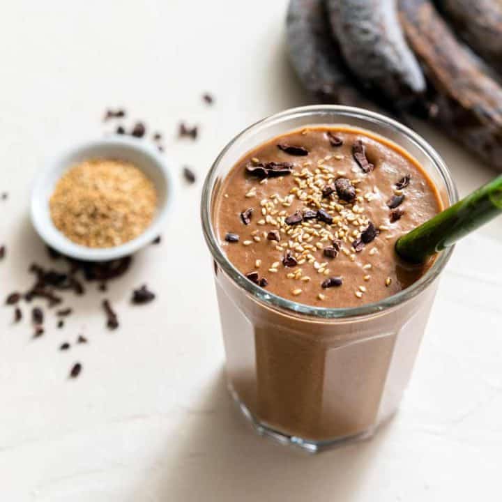 One large glass filled with chocolate sesame smoothie, with seeds, nibs, and bananas on the side