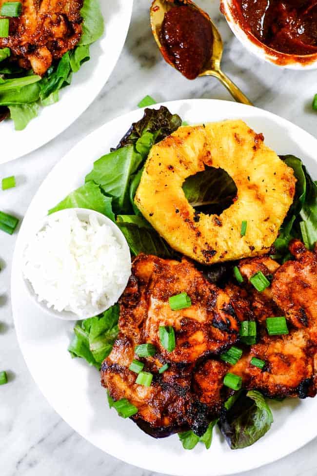 Grilled pineapple chicken on a plate of greens with extra bbq sauce and a side of white rice.