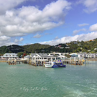 Paihia Wharf in the Bay of Islands, New Zealand – charming white waterfront buildings, cafés, and shops along the harbour, with a blue cruise boat docked and green hills rising behind under a sunny sky. Paihia waterfront, Bay of Islands: white colonial buildings and wharf with cafés, blue boat in turquoise harbour, green hills and cloudy blue sky, New Zealand.