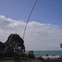 The iconic Wind Wand kinetic sculpture swaying in the breeze along the New Plymouth foreshore. The Len Lye Wind Wand kinetic sculpture, a tall red fiberglass pole with a glowing glass orb on top, bending against a blue sky on the New Plymouth Coastal Walkway.