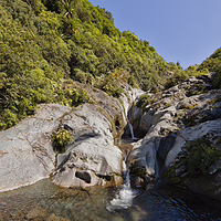 The pristine, crystal-clear mountain water of Wilkies Pools. A close-up view of clear, shallow alpine water flowing over smooth grey rocks into natural mountain pools at Wilkies Pools in New Plymouth, surrounded by lush green native forest under a bright blue sky.