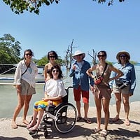 Group of travelers, including a person in a wheelchair, enjoying a sunny day by the water. Exploring the Philippines with a diverse group by the beach.