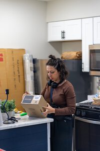 Alternate text: Woman with headphones unpacking a delivery box in contemporary kitchen with white cabinets.