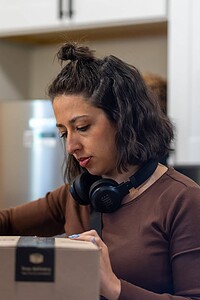 Close-up of focused woman packaging products in kitchen, wearing headphones, with a black shirt and dark hair.