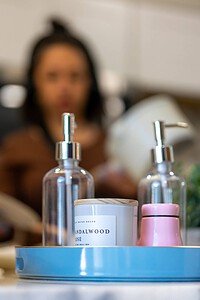 Close-up of stylish soap dispensers, candles, and decor on kitchen countertop with blurred woman in background.