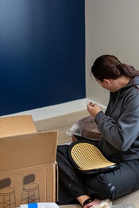 Woman working on pottery or craft project sitting on floor, surrounded by art supplies and a cardboard box, in a cozy studio setting.