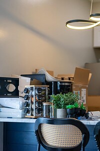 A messy kitchen workspace with unpacked boxes, cleaning tools, and miscellaneous items on the counter.