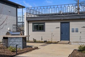 A small, contemporary outdoor space with a grill and a beige building featuring a blue door and window.