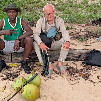 Two men sitting together on a sandy beach next to several whole green coconuts.
