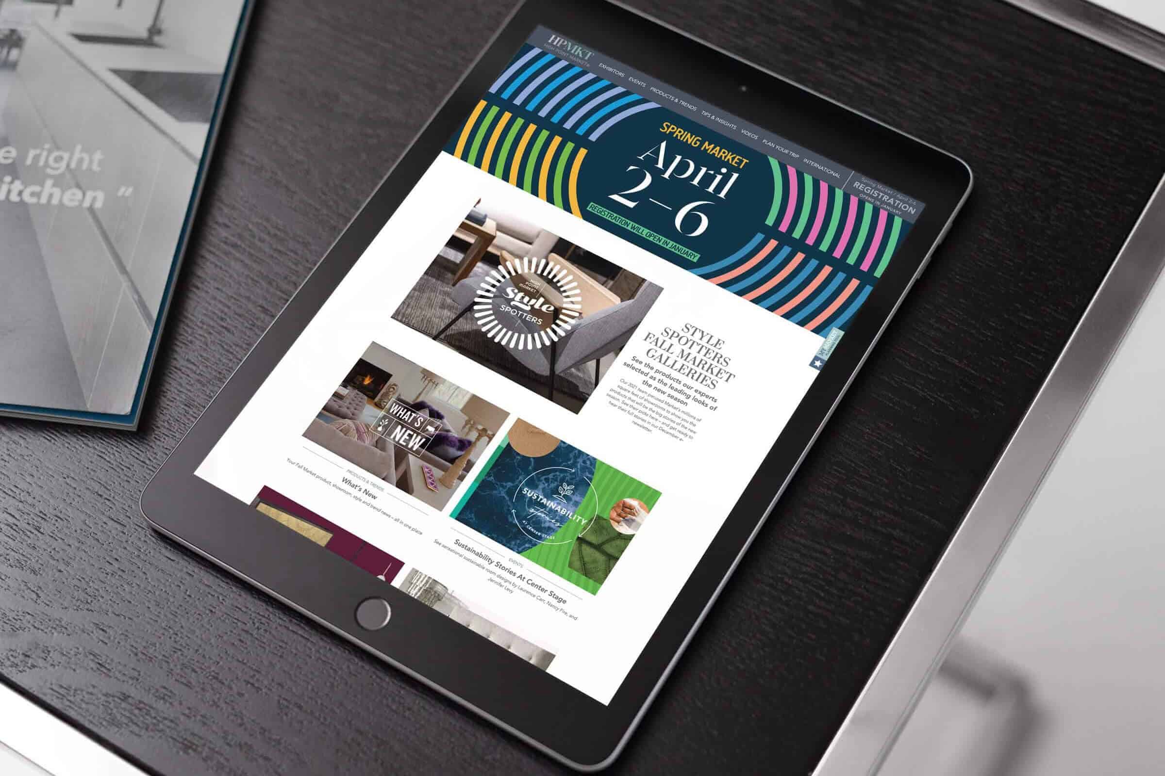 An ipad on a desk that shows the High Point Market home screen. It displays simple article blocks that contain colorful photographs