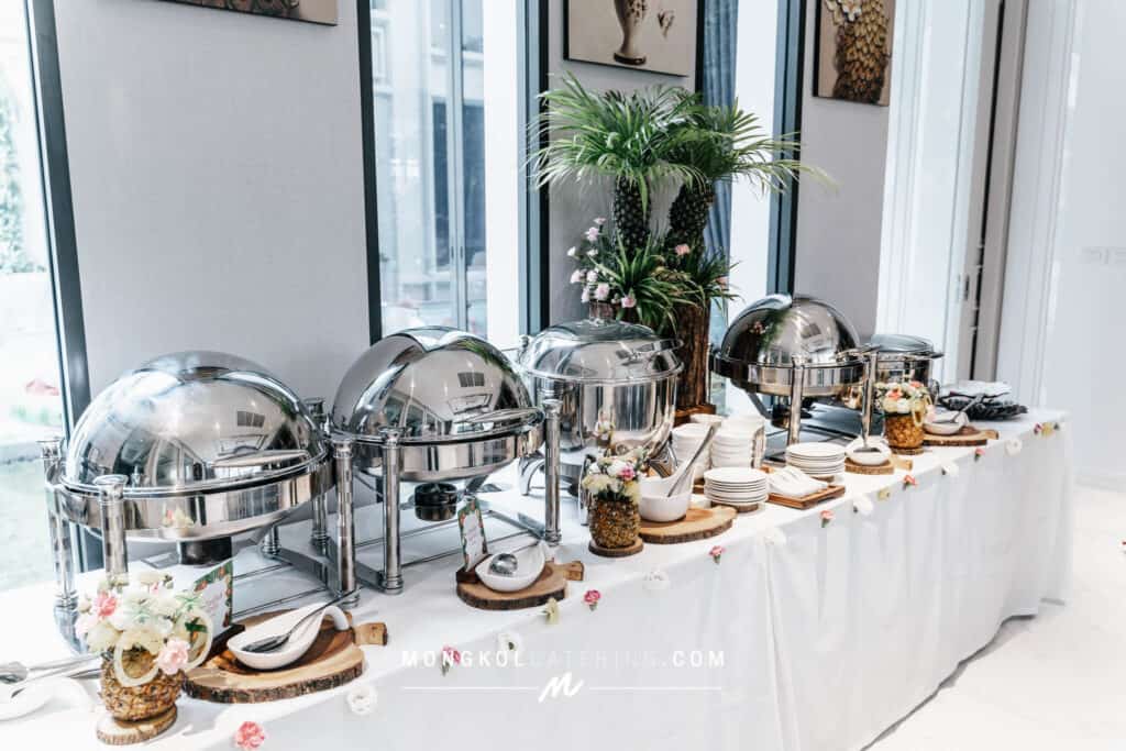 Shuttle buffet setup with chafing dishes, floral decorations, and elegant tableware in a bright event space in Thailand.