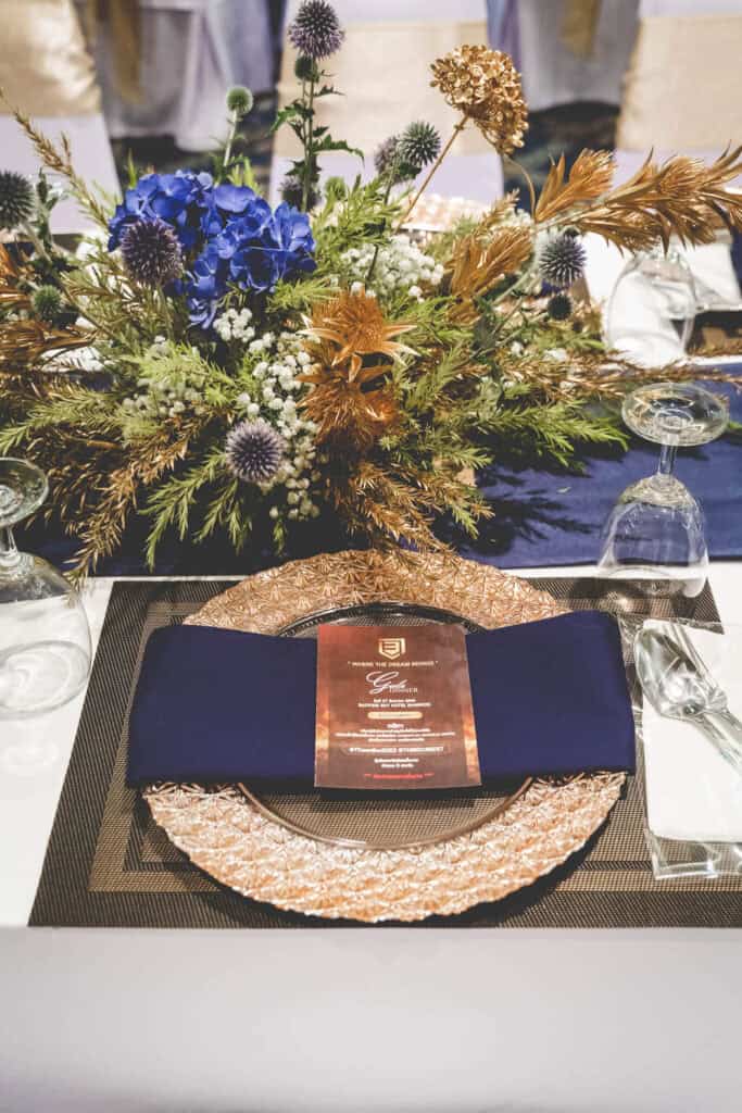Colorful floral table centerpiece at a Thai event, featuring blue, white, and gold flowers with elegant tableware.