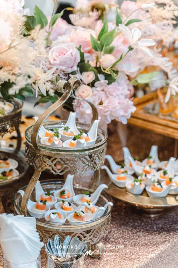 Elegant appetizer display with small seafood bites on a silver stand, surrounded by pink flowers and lush decor in Thailand.