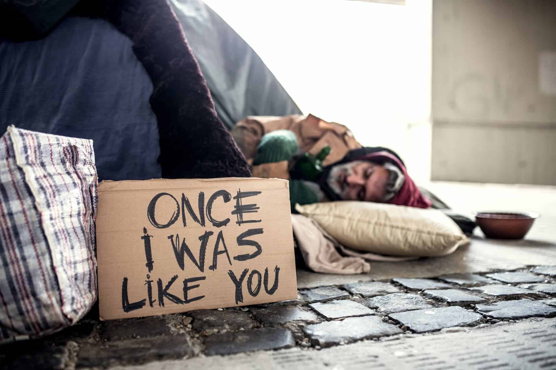 A homeless man lies under a bridge in Tampa, wrapped in worn clothing, holding a cardboard sign that reads 'ONCE I WAS LIKE YOU,' highlighting the realities of homelessness.
