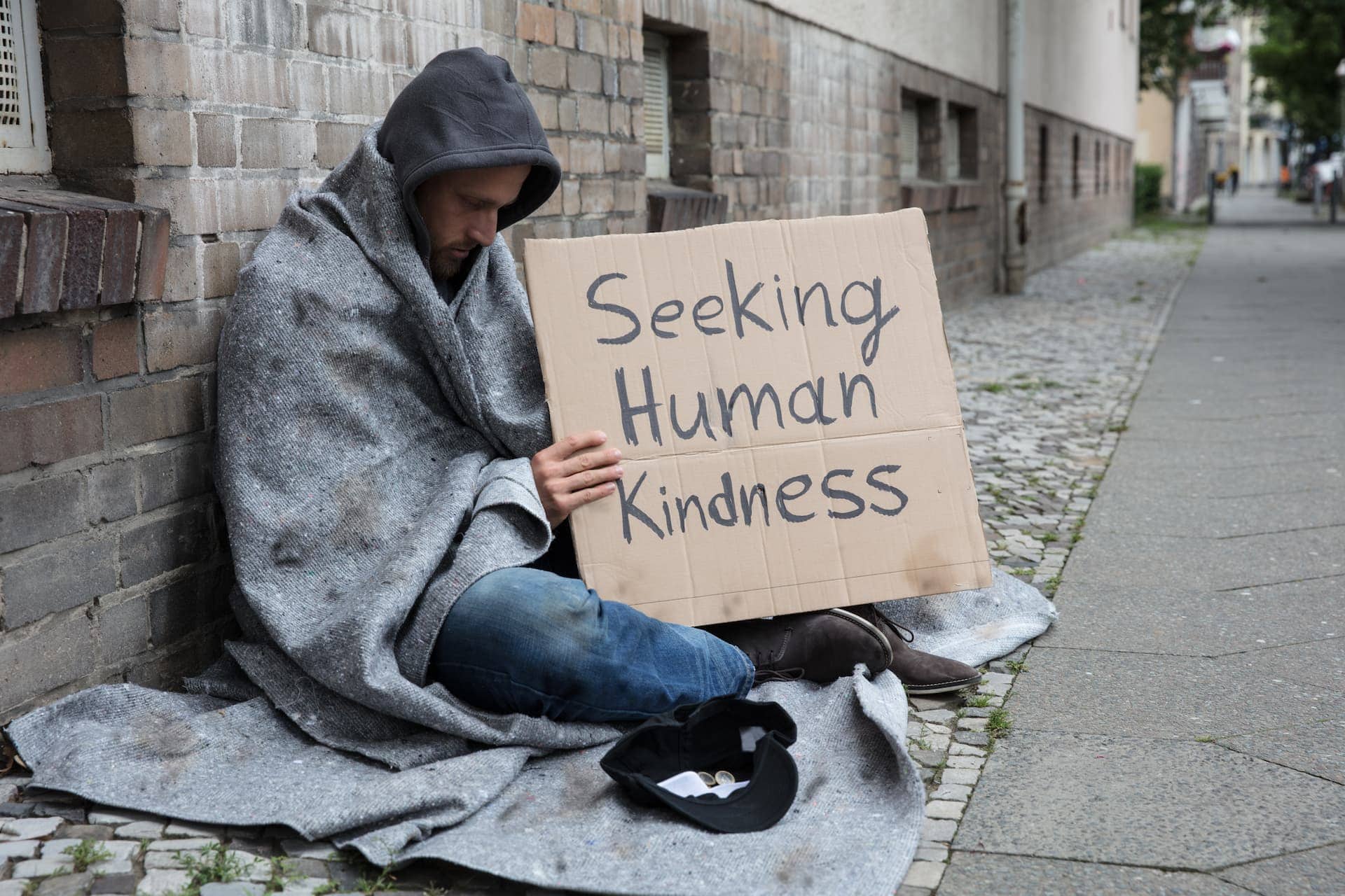 A homeless man sits wrapped in a blanket on a Tampa city sidewalk, holding a sign that reads 'Seeking Human Kindness'.