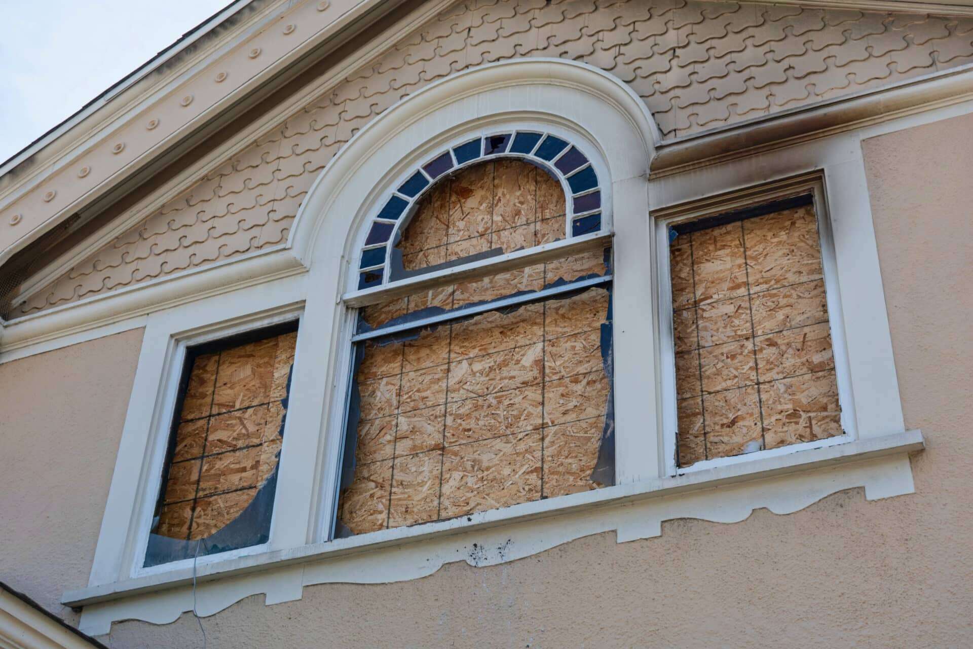Home window boarded up with plywood sheets. House fire walls and eves charred. Plywood shutters prevent unauthorized access by squatters, looters to damaged unused, vacant, or abandoned property.