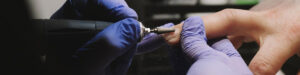 A person wearing blue gloves uses a nail drill on another's fingernails, flakes of nail visible as they are filed. In a well-lit setting with proper salon ventilation, the process takes place against a blurred background, highlighting the focused nail grooming. Read more at Aerovex Systems (aerovexsystems.com)