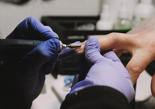 Image of a nail technician using an electric file on a client's fingernail. Nail dust is seen rising up from the tool leading to a discussion on why is proper salon ventilation in the nail industry so crucial.