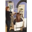 A stylist with long reddish hair finishes a keratin treatment on a client's long brown hair in a salon decorated with blue walls and white trim. The client is seated in a white chair, wearing a black cape, facing a mirror. Behind them is The Original Chemical Source Capture System™. A Source Capture System is the first step to implementing 3-Zone Protection in hair salons. Read more at Aerovex Systems (aerovexsystems.com)