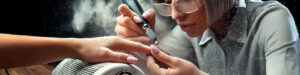 A nail technician wearing glasses shapes a client’s nails with an electric nail file, using a nail dust collector that is releasing harmful nail dust into the air, illustrating the question, do cheap nail dust collectors work? The client’s nails are painted light pink. The scene is close-up and focused on the hands and tools used during the manicure. Read more at Aerovex Systems (aerovexsystems.com)