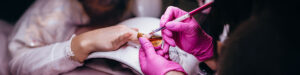 A close-up of a person getting a gel nail manicure. Another person wearing bright pink gloves carefully shapes a nail with a brush, while the client’s hand rests on a white cushion. The focus is on the hand and nail work which indicates airborne VOCs for a discussion on cleaner nail salon air, with blurred background details. Read more at Aerovex Systems (aerovexsystems.com)