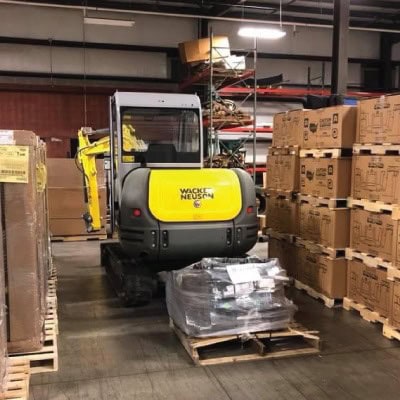 Small yellow and black compact loader in warehouse with stacked boxes.