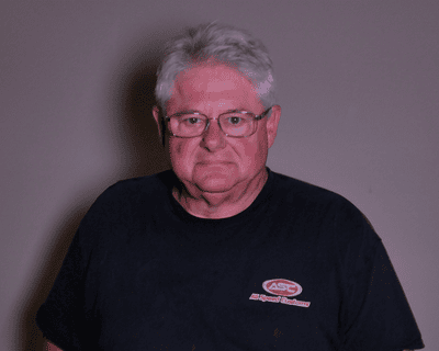 Wally Ochs, an older man with glasses and gray hair, wearing a black T-shirt with All Speed Customs.