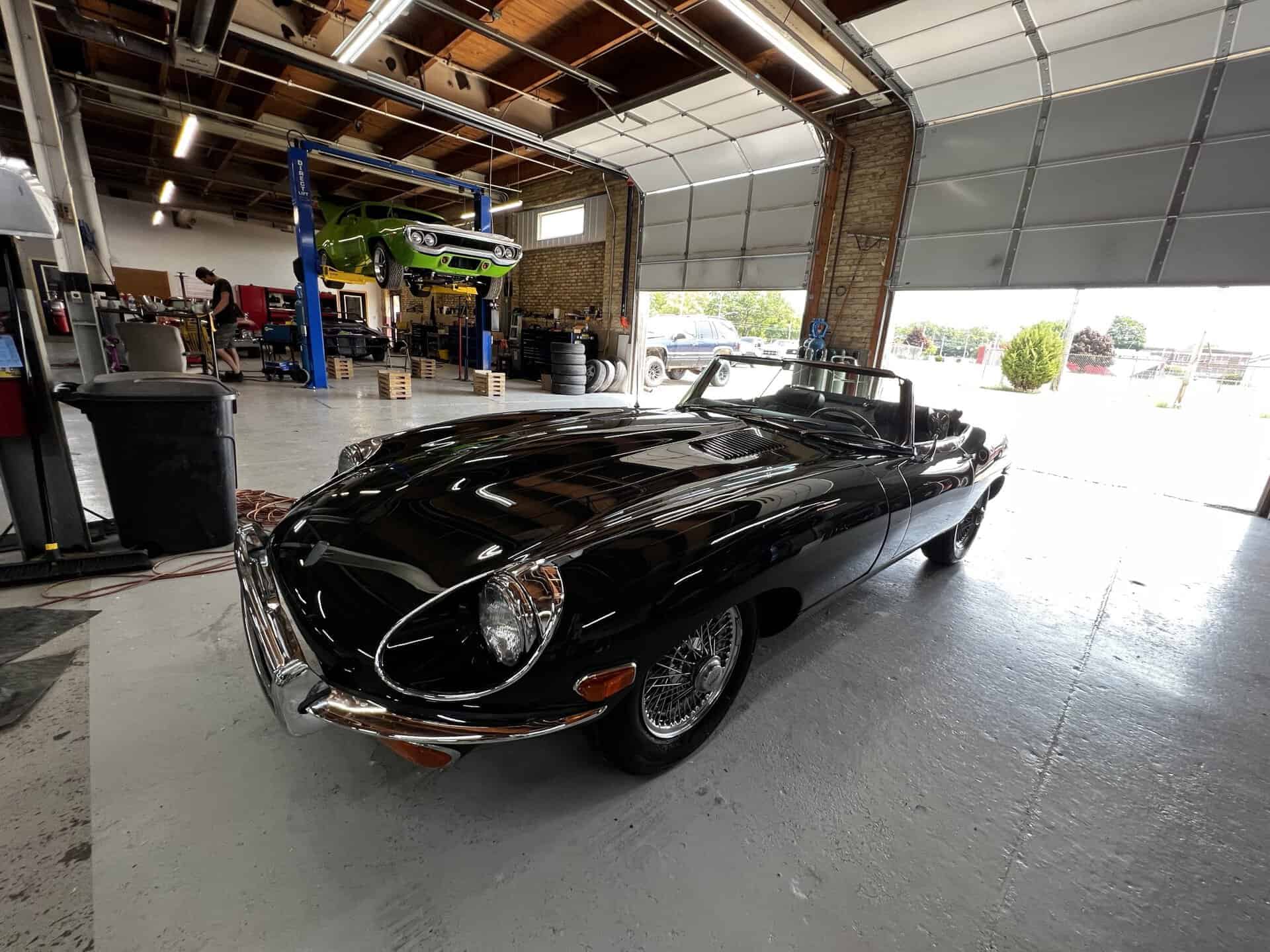 Black 1970 Jaguar E-Type in a garage with car lift and tools.
