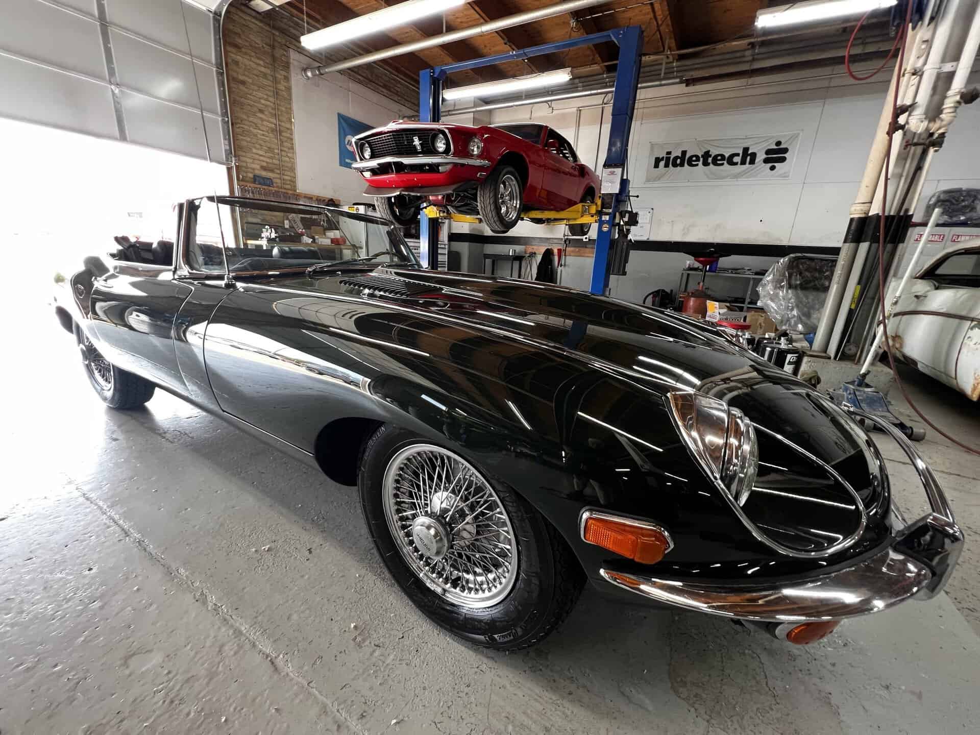 Black 1970 Jaguar E-Type in workshop with red car on lift.