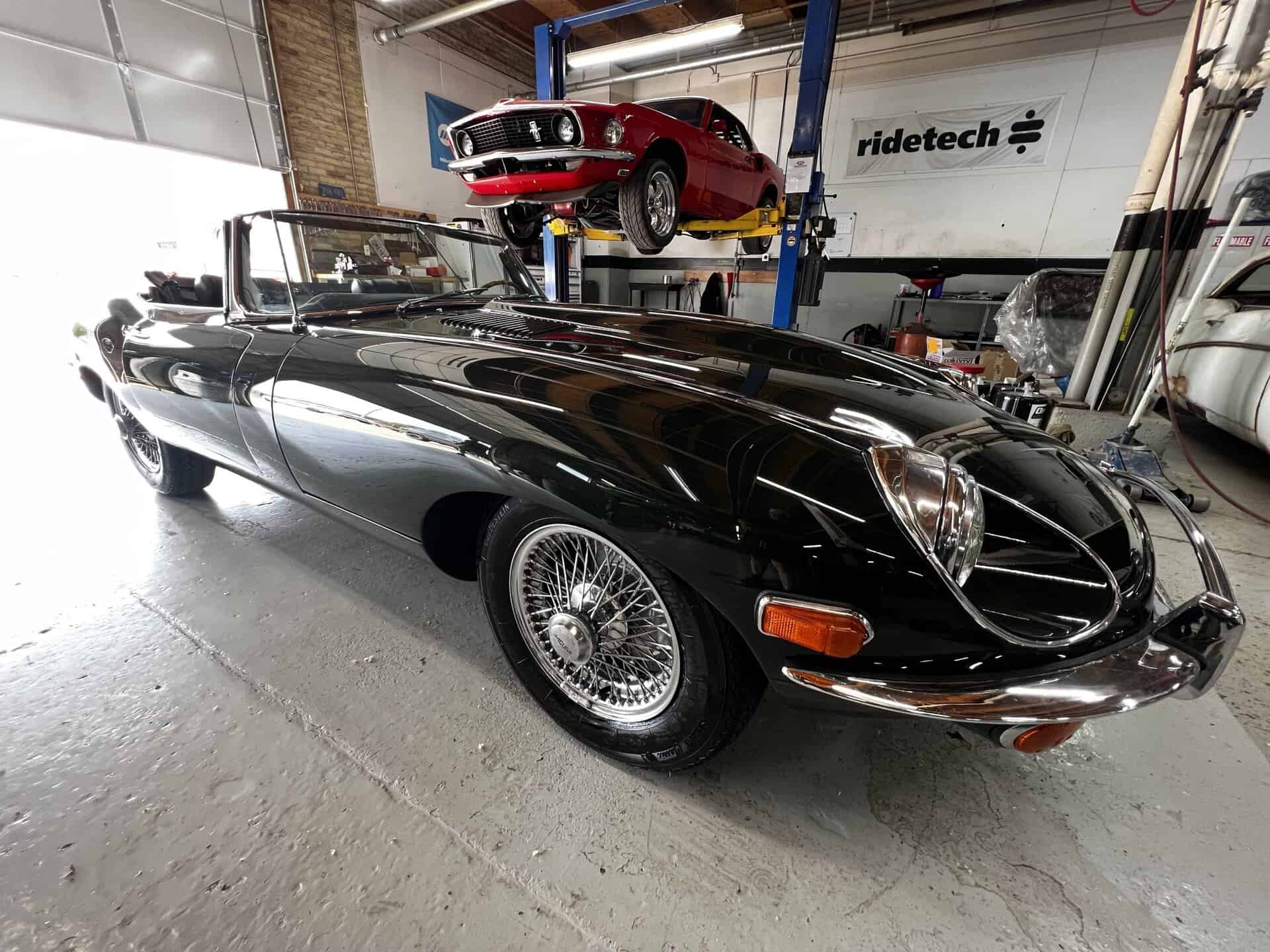 Black 1970 Jaguar E-Type in a garage with a red car on a lift.