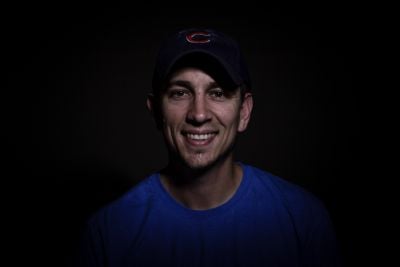 Smiling man wearing a baseball cap and blue shirt against dark background.