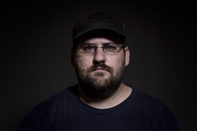 Close-up portrait of a man with glasses, beard, and dark background.