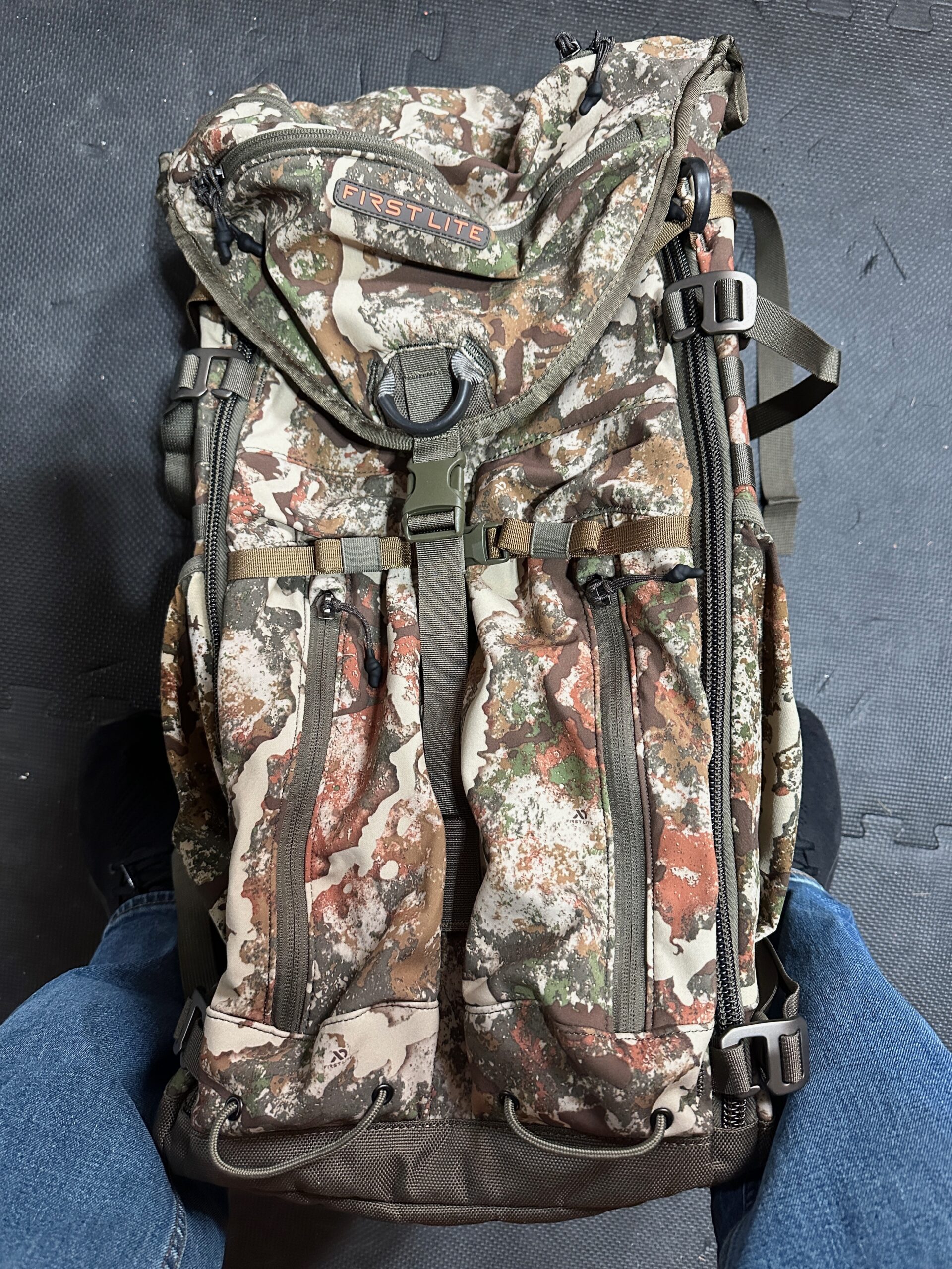 Front of First Lite Transfer Pack showing zippered storage compartments, compression straps, and mobile treestand attachment points