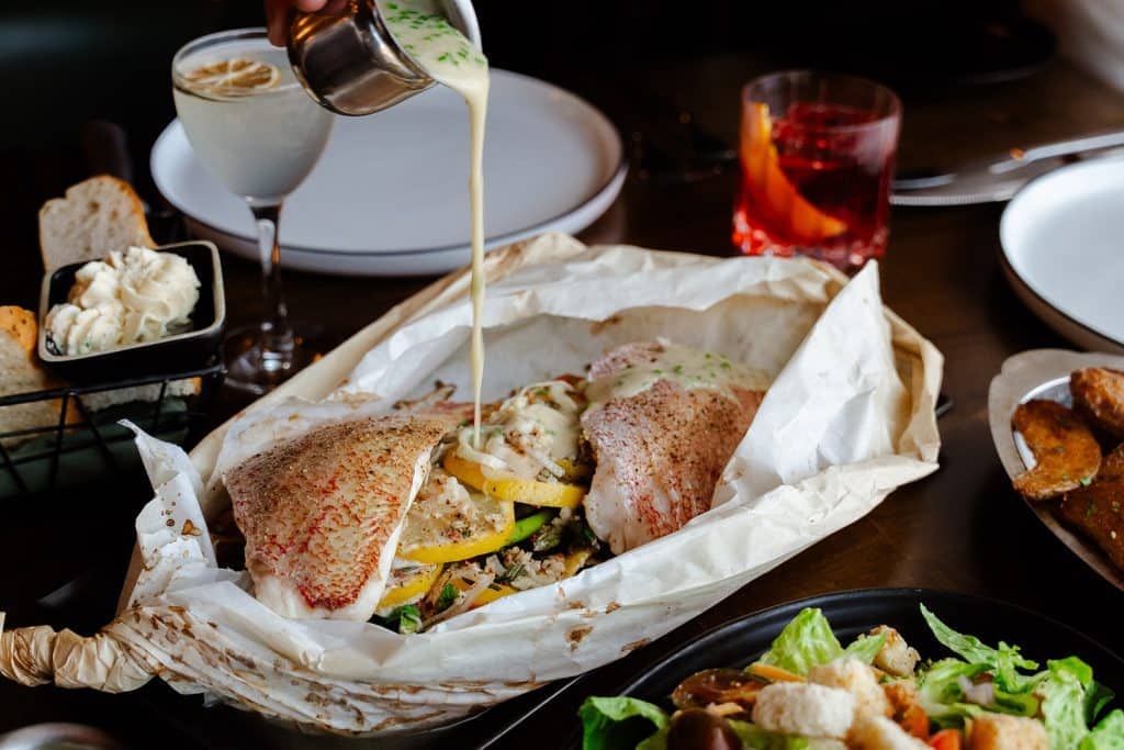 Fresh fish fillet being poured with creamy sauce, served with vegetables and lemon wedge