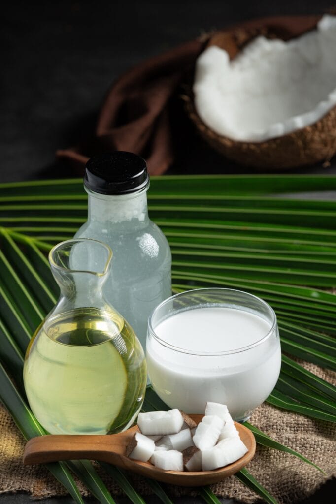 Coconut oil in a glass carafe next to other coconut products and pieces of fresh coconut