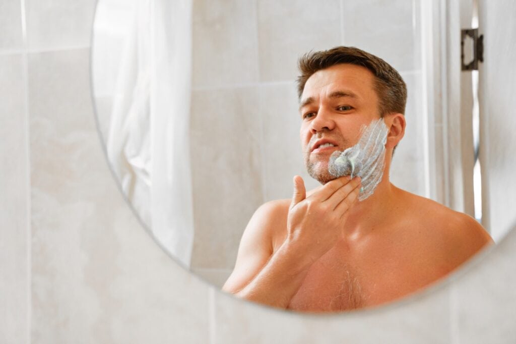 Man working shaving cream into his cheek with his fingers, seen in a round bathroom mirror