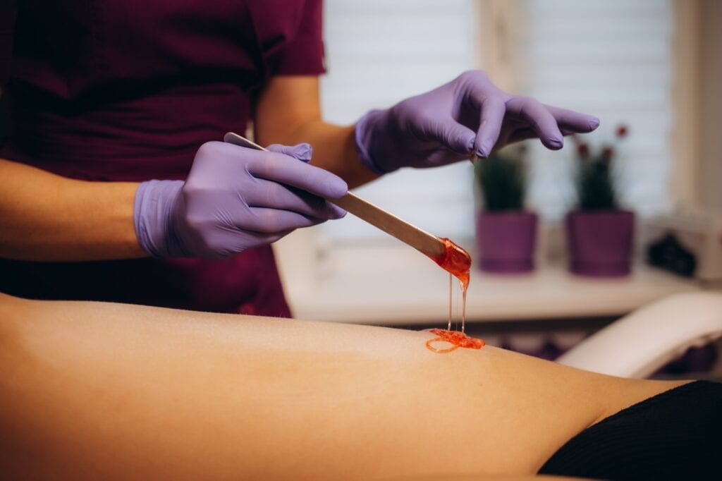 Therapist in purple gloves applying sugar paste to a client's leg with a wooden stick