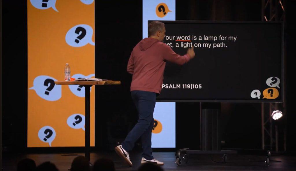 Preacher presenting sermon illustration on stage with question marks background.