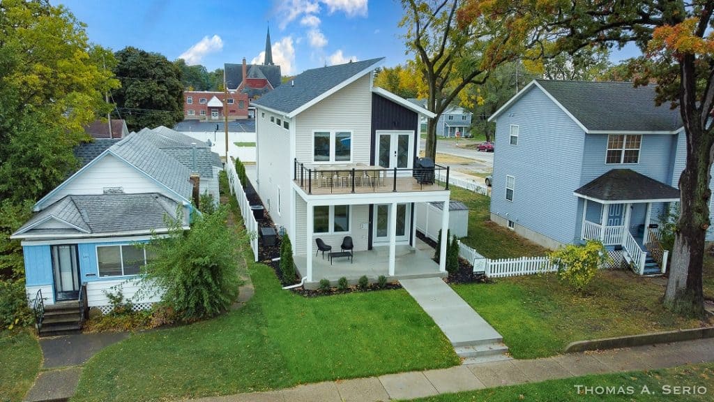 A modern two-story custom home with a large front porch and balcony, surrounded by greenery.