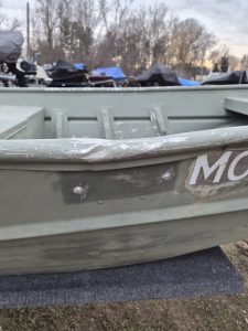 Close-up of a weathered boat hull showing minor damage and repairs, with boat motors and outdoor storage in the background.